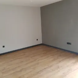 Wykończone wnętrze pokoju z panelami podłogowymi w odcieniach drewna, szarą ścianą z gładzią i białą ścianą z widocznymi gniazdkami oraz czarnymi listwami przypodłogowymi.