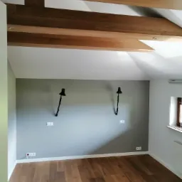 Wnętrze pokoju w Warszawie po remoncie: drewniana podłoga, szara ściana z dwoma czarnymi lampami, drewniane belki na suficie.