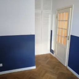 Ściana pomalowana do połowy na granatowo, górna część biała, parkiet w jodełkę, biała szafa z żaluzjowymi drzwiami i częściowo otwarte białe drzwi z szybkami.