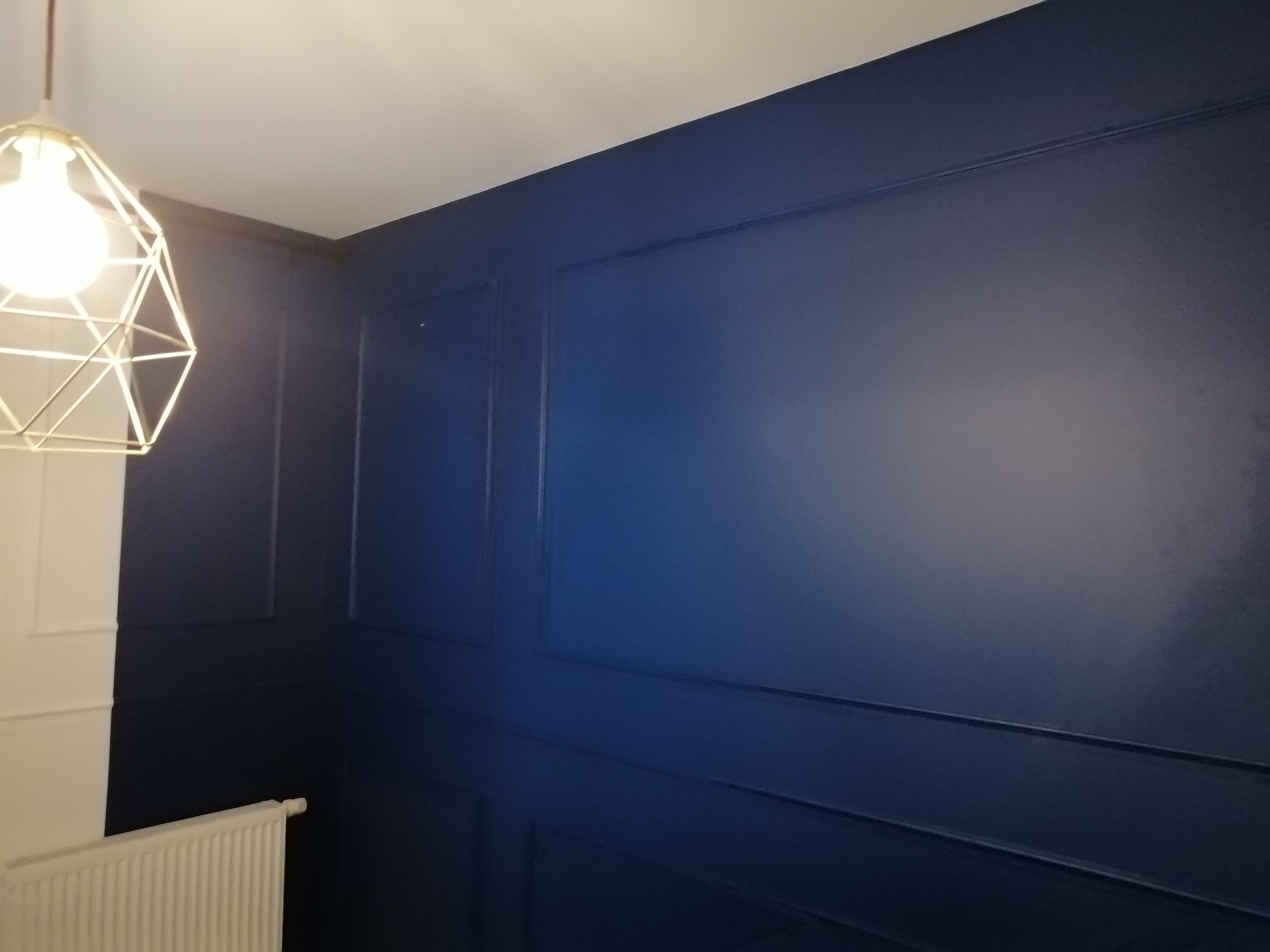 Granatowa ściana z dekoracyjnymi listwami w stylu angielskim, oświetlona nowoczesną lampą wiszącą z geometrycznym kloszem, widoczny biały grzejnik.