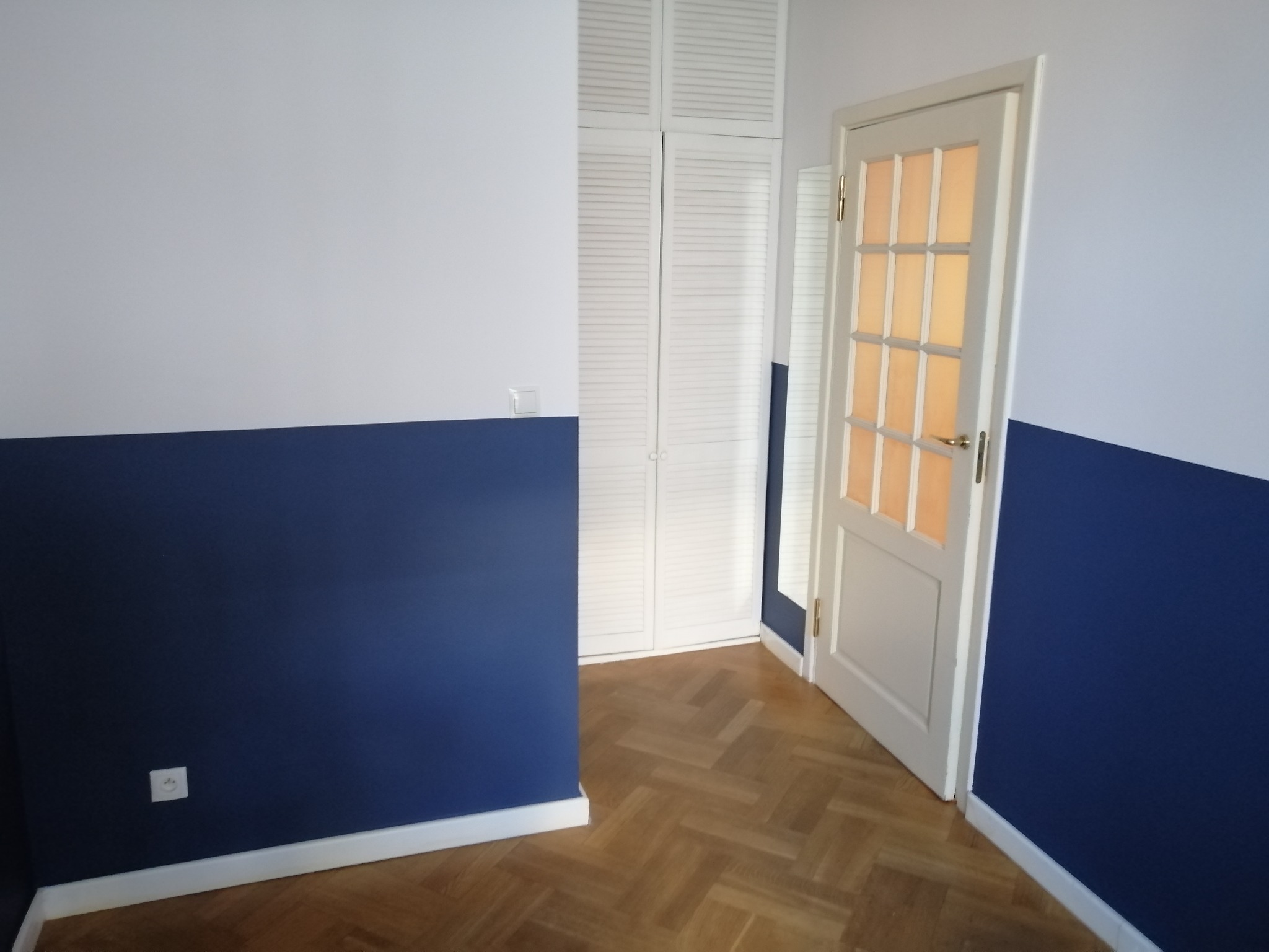 Ściana pomalowana do połowy na granatowo, górna część biała, parkiet w jodełkę, biała szafa z żaluzjowymi drzwiami i częściowo otwarte białe drzwi z szybkami.