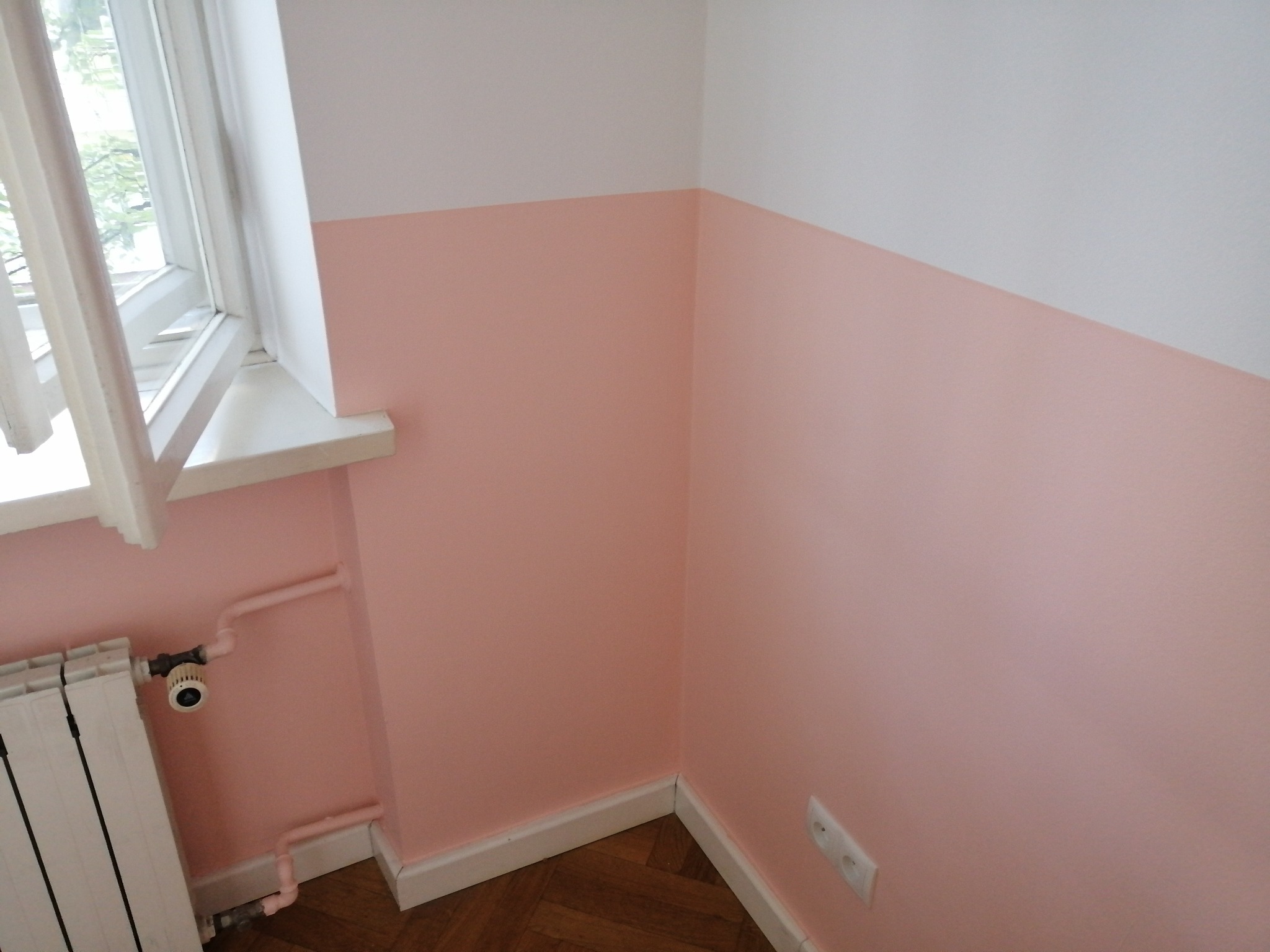 Pomieszczenie po malowaniu ścian na dwa kolory: biały u góry i łososiowy na dole, widoczny biały grzejnik i drewniana podłoga parkietowa.