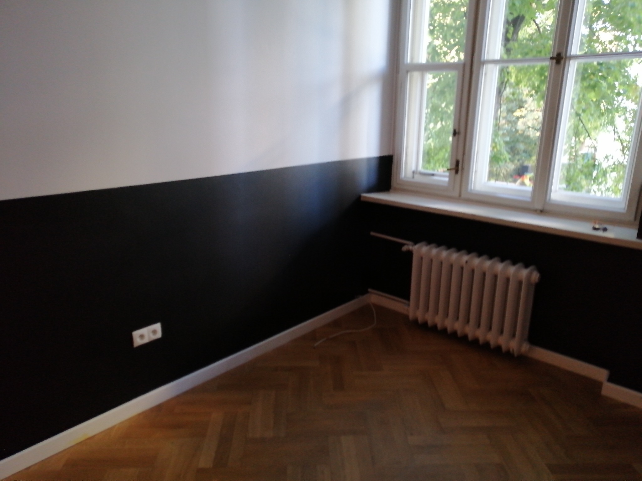 Pokój po remoncie: ściany pomalowane do połowy na biało i czarno, parkiet w jodełkę, biały grzejnik pod oknem.