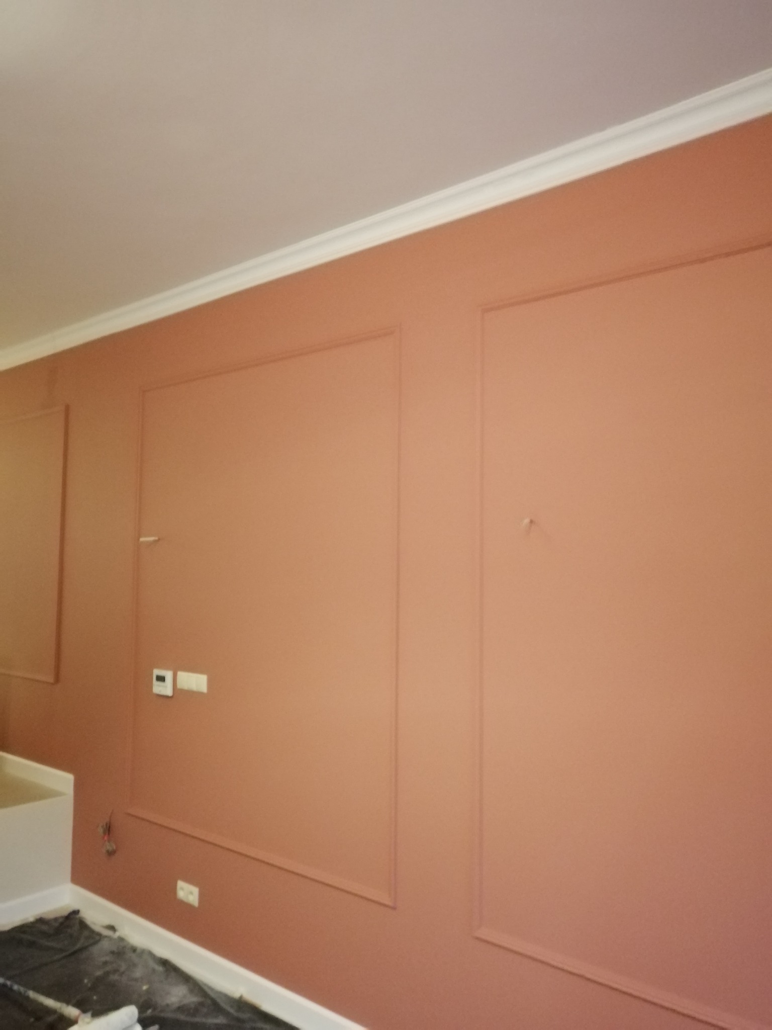 Ściana pomalowana na kolor terakoty z dekoracyjnymi ramkami i białą listwą przysufitową, widoczne gniazdka elektryczne i wystające kable.