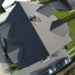 Widok z drona na dom z ciemnoszarą dachówką, z trzema lukarnami i kominem z anteną satelitarną, otoczony zadbanym ogrodem i dwoma samochodami na podjeździe.