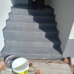 Daniel Majewski - Wykończone schody wejściowe z szarych płytek, widoczne dwa wiadra z zaprawą i narzędzia. Ujęcie z dołu, widać fragment elewacji i drzwi wejściowe.