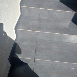 Daniel Majewski - Wykończone schody z szarych płytek z jasnymi fugami, przylegające do białej ściany o wyraźnej fakturze. Widoczne cienie padające na schody.