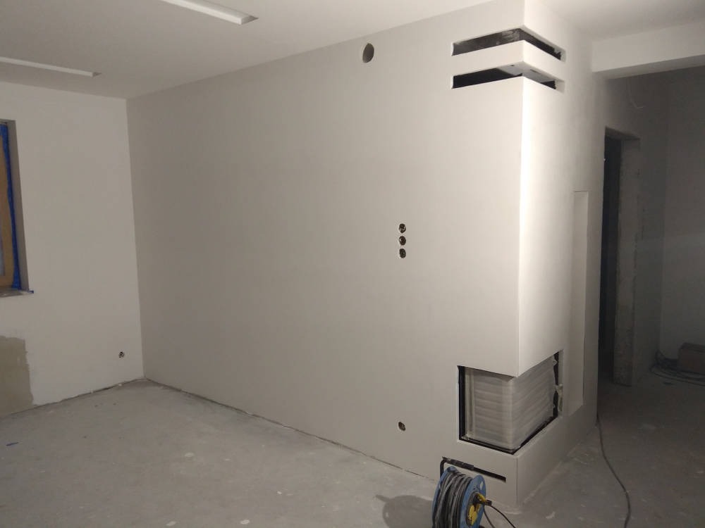 Wnętrze po szpachlowaniu ścian na gładko, z widocznym narożnikiem obudowy kominka z otworami wentylacyjnymi i przygotowanymi punktami elektrycznymi.