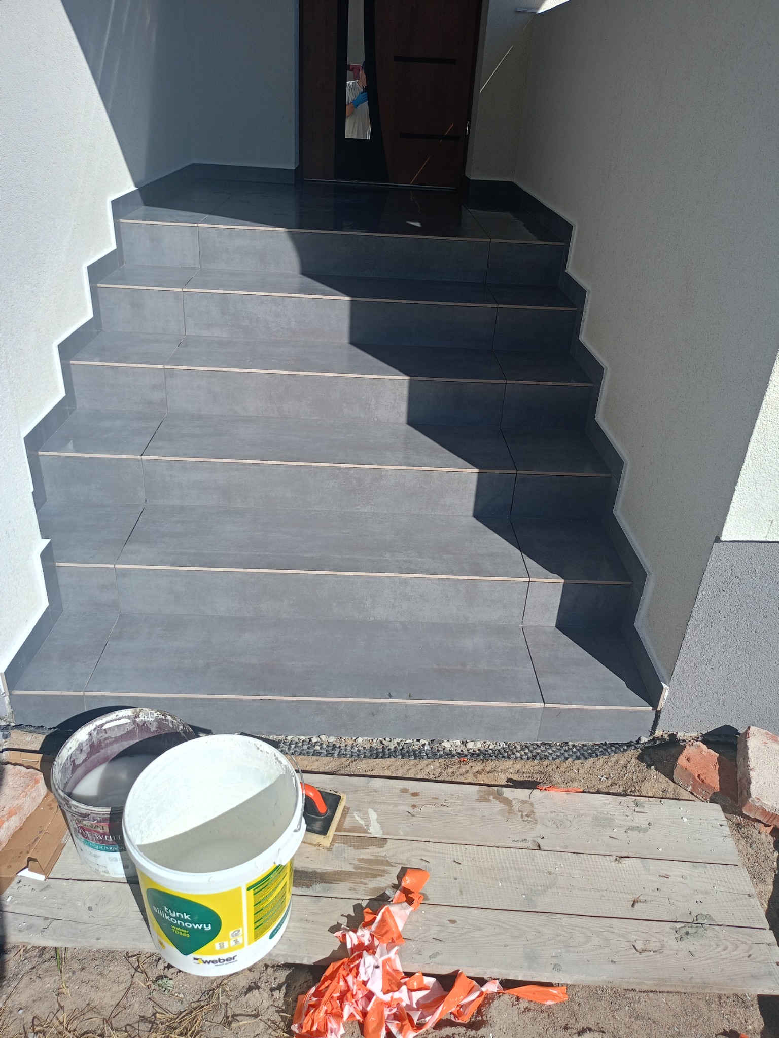 Wykończone schody wejściowe z szarych płytek, widoczne dwa wiadra z zaprawą i narzędzia. Ujęcie z dołu, widać fragment elewacji i drzwi wejściowe.