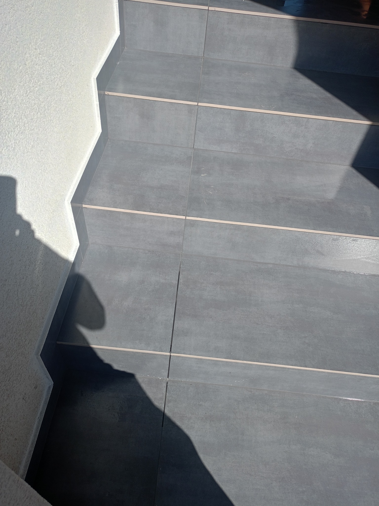 Wykończone schody z szarych płytek z jasnymi fugami, przylegające do białej ściany o wyraźnej fakturze. Widoczne cienie padające na schody.