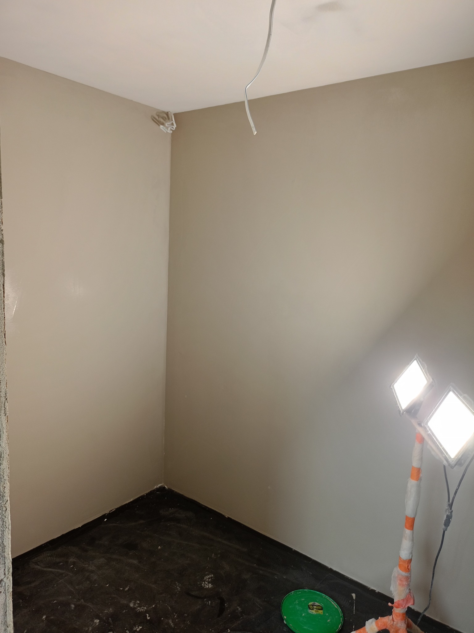 Wnętrze po szpachlowaniu, ściany w beżowym odcieniu, zabezpieczona podłoga, lampa budowlana i widoczne ślady po pracy.