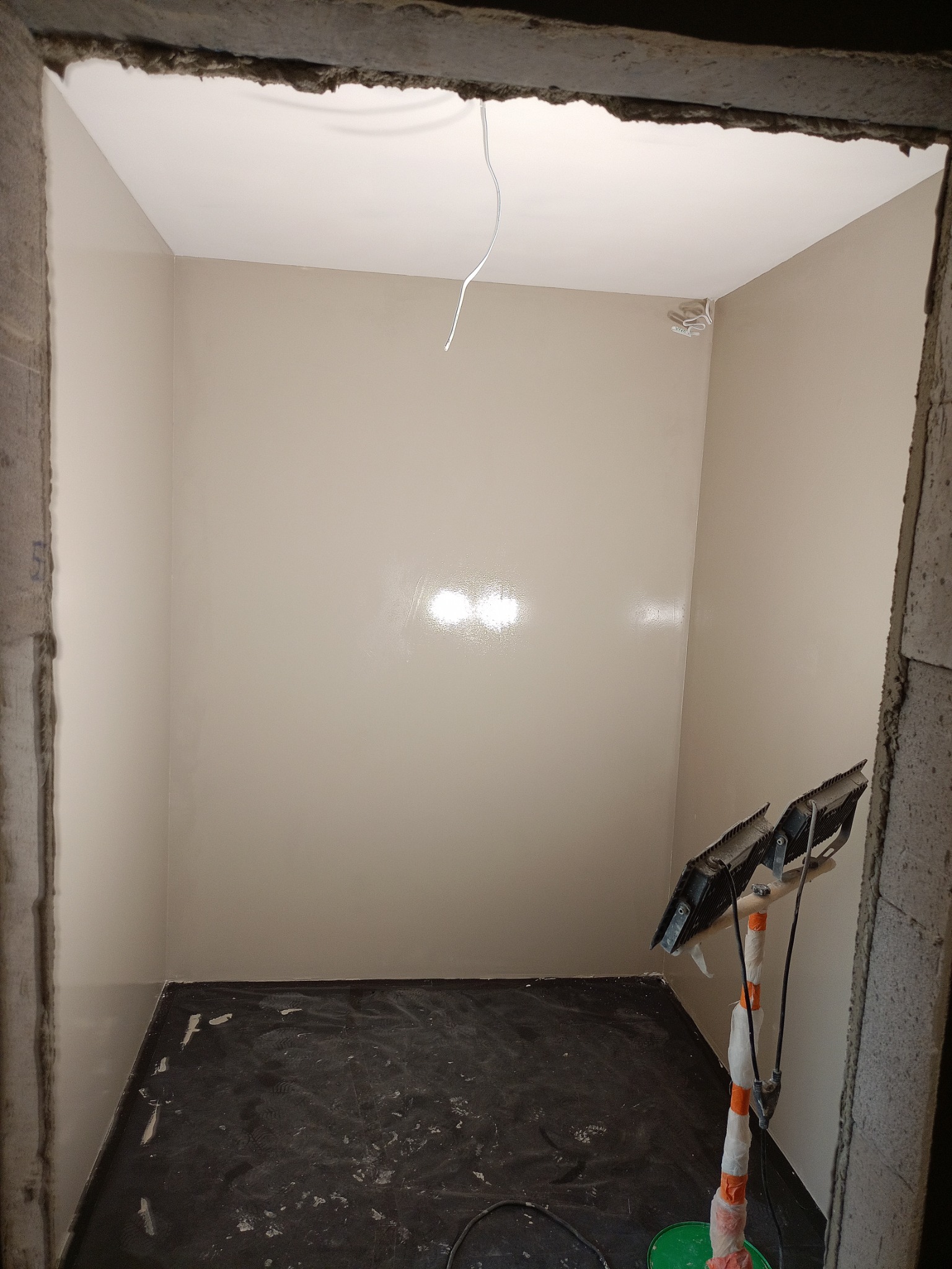 Wnętrze po szpachlowaniu: gładkie, beżowe ściany w surowym otoczeniu, widoczne narzędzia pracy i zabezpieczona podłoga. Oświetlenie halogenowe.
