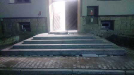 Zewnętrzne schody wejściowe wyłożone szarymi płytkami, prowadzące do otwartych drzwi budynku z kamienną elewacją i małymi oknami piwnicznymi.