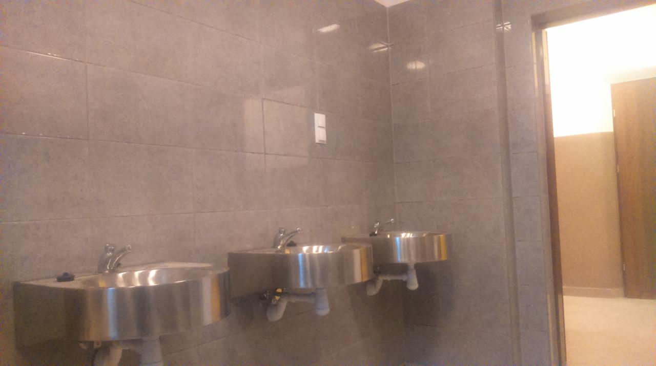 Wykończona łazienka publiczna z trzema metalowymi umywalkami zamontowanymi na szarej, prostokątnej ścianie z płytek, widoczny fragment otwartych drzwi do kolejnej części łazienki.