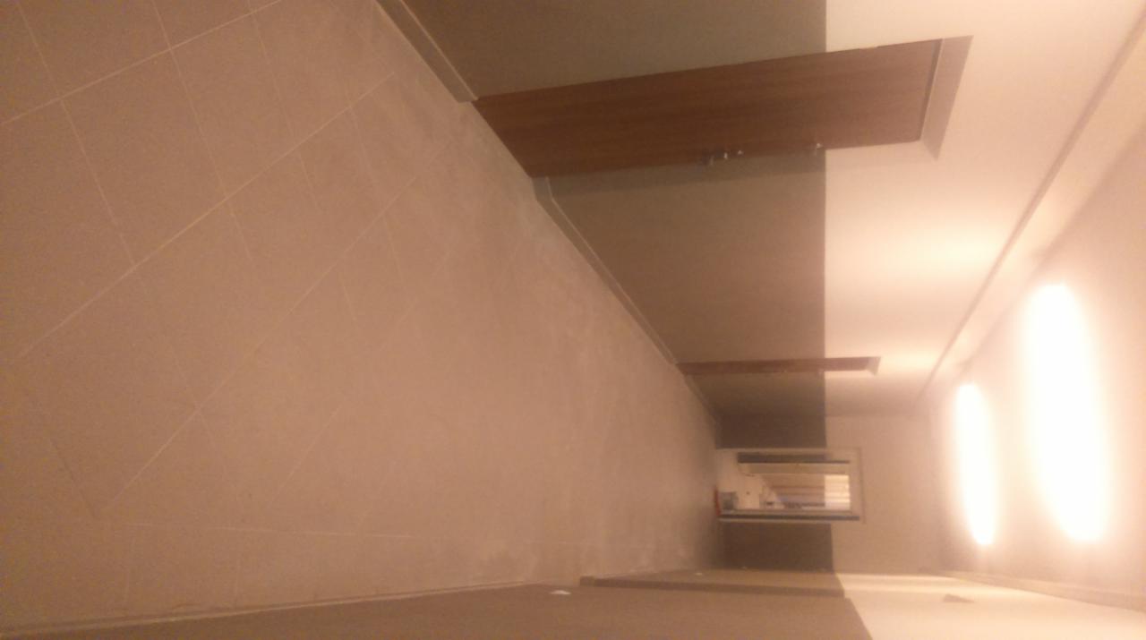 Ściana wyłożona beżowymi płytkami ceramicznymi z widocznymi fugami, fragment sufitu z drewnopodobną listwą dekoracyjną i jasnym oświetleniem, wnętrze w trakcie lub po remoncie.