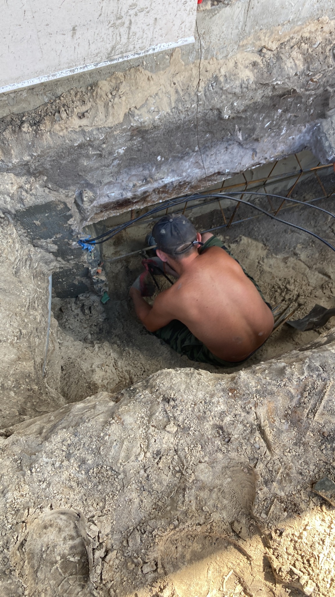 Pracownik w wykopie fundamentowym w Chorzowie, montuje zbrojenie; widoczne przewody i narzędzia.