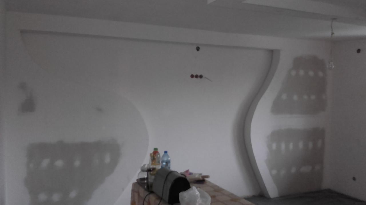 Niedokończona ściana z dekoracyjnymi, gipsowymi elementami w kształcie fal, widoczne ślady szpachlowania i otwory na gniazdka elektryczne, na pierwszym planie blat z butelkami wody i narzędziami.