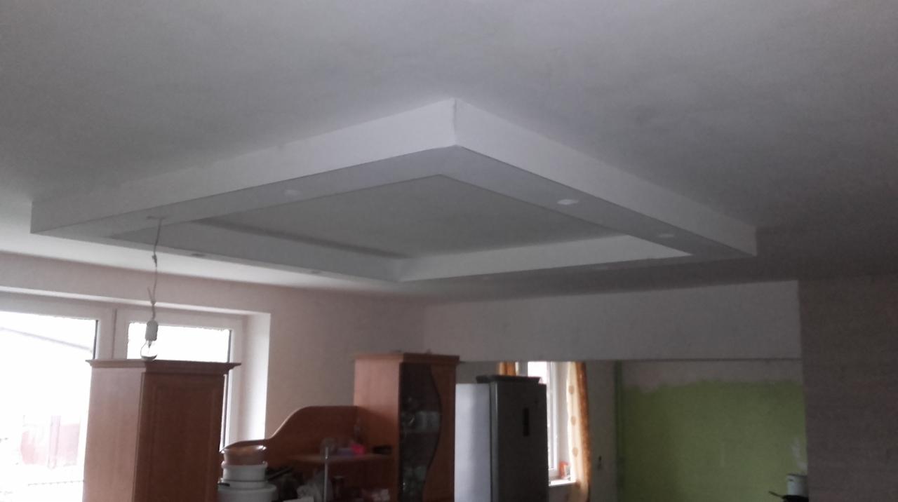 Wykończony sufit podwieszany z oświetleniem punktowym, widok na częściowo umeblowane wnętrze z oknem i elementami w trakcie prac.