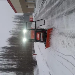 Pomarańczowy traktor z pługiem odśnieżającym, z włączonymi światłami, usuwa śnieg z drogi w pochmurny dzień. Ślady opon widoczne na śniegu. W tle budynek i drzewa bez liści.