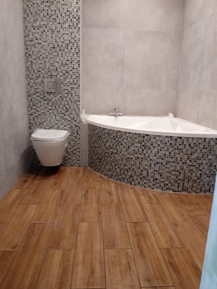 Nowoczesna łazienka z narożną wanną obudowaną mozaiką, drewnopodobną podłogą i toaletą podtynkową na tle ściany z mozaiki. Na wannie widoczny dozownik mydła.