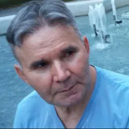 Portret mężczyzny z siwiejącymi włosami na tle fontanny z figurami, ubrany w błękitną koszulkę.