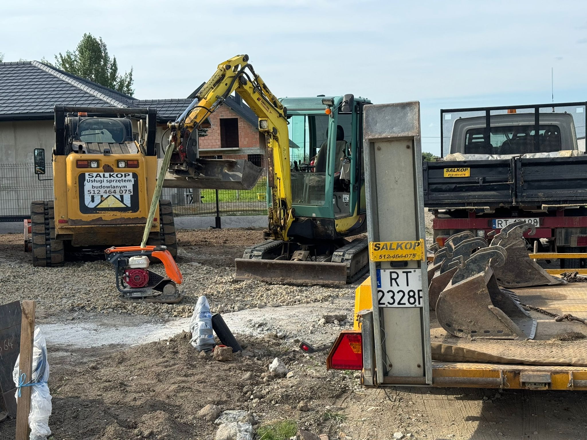Żółta miniładowarka i koparka na placu budowy z przyczepą pełną łyżek. W tle dom w trakcie budowy. Widoczny zagęszczarka gruntu i tablica z nazwą firmy.
