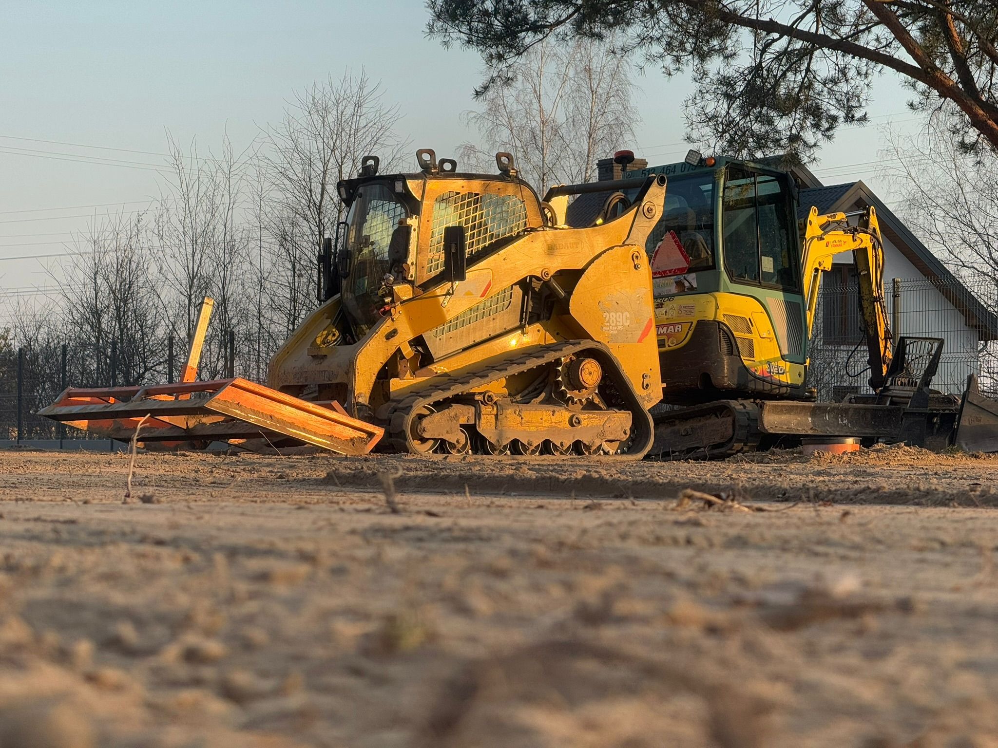 Żółta miniładowarka gąsienicowa i koparka na placu budowy, z widocznym domem w tle. Sprzęt budowlany przygotowany do prac ziemnych.
