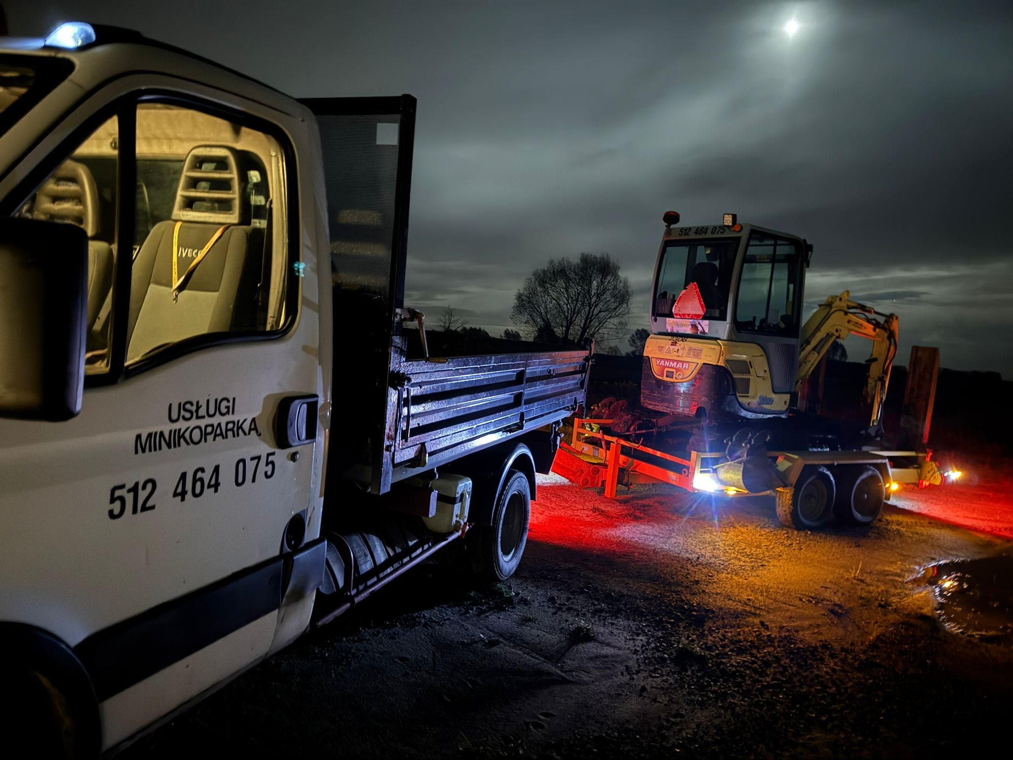 Nocny transport minikoparki Yanmar na lawecie, załadowanej na skrzynię samochodu dostawczego z napisem 'Usługi Minikoparką' i numerem telefonu, oświetlonej światłami.