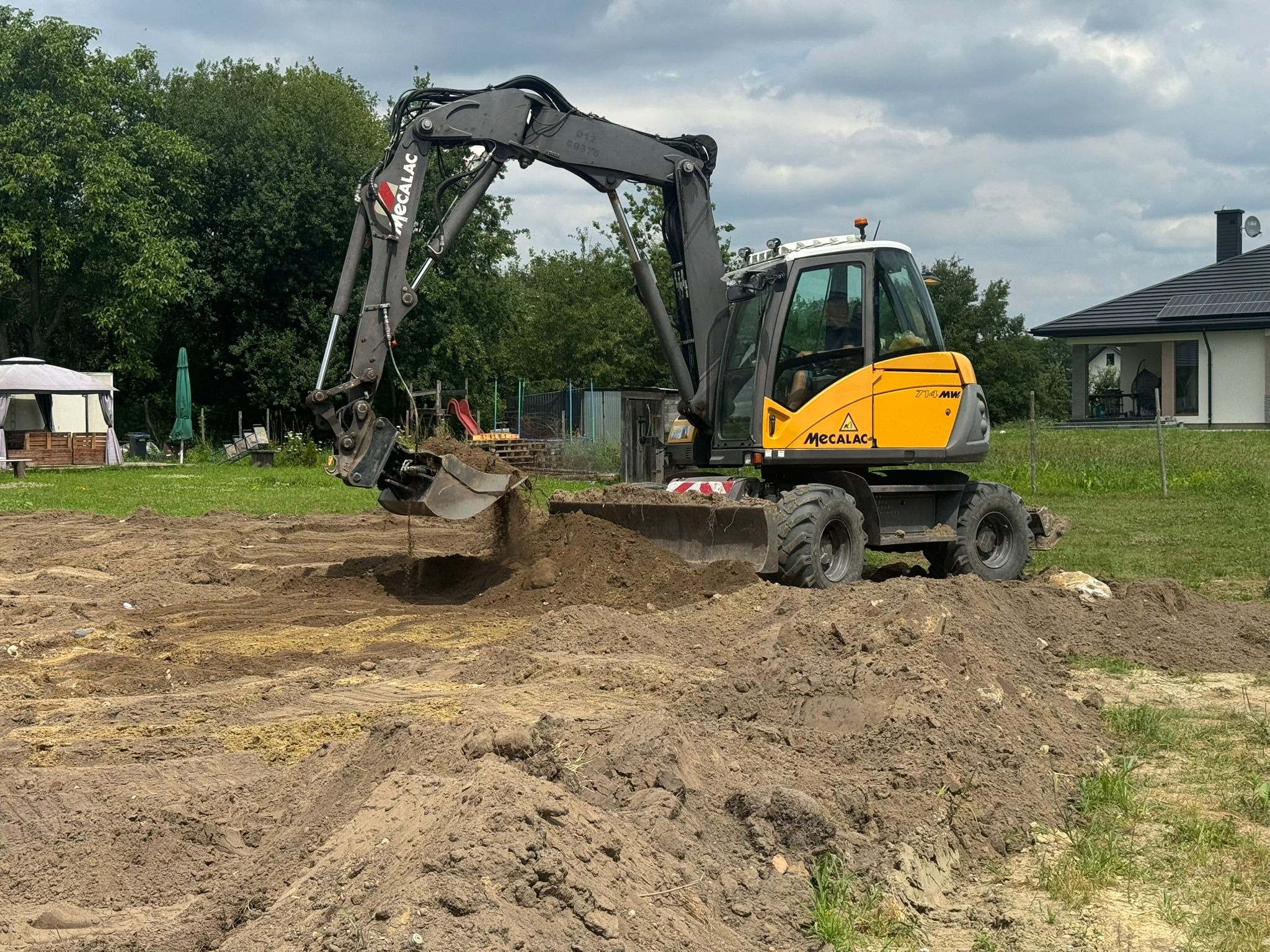 Żółto-szara koparka Mecalac w trakcie prac ziemnych na działce z widocznym domem w tle. Koparka zagłębia się w ziemię, tworząc wykop.