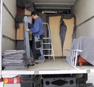 Wnętrze samochodu dostawczego wypełnione meblami zabezpieczonymi szarymi kocami, mężczyzna w trakcie wnoszenia składanej drabiny aluminiowej, sterta koców na podłodze, biała drabina oparta o ścianę.