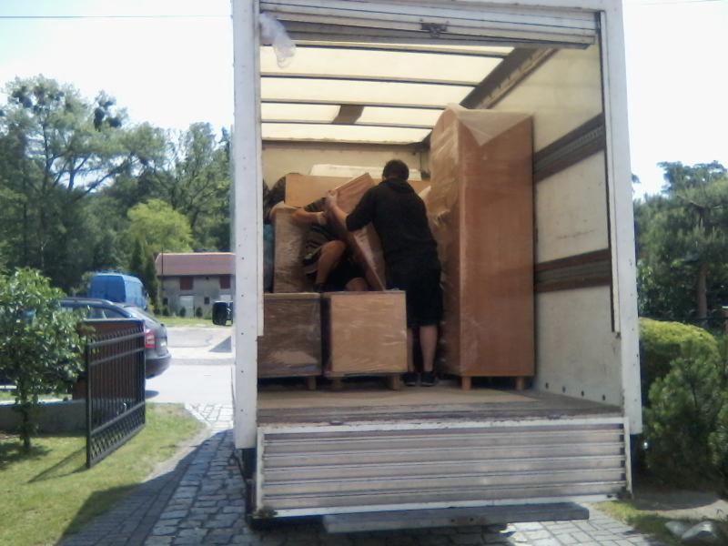 Mężczyźni układają meble owinięte folią wewnątrz białej furgonetki z otwartymi drzwiami, widok z zewnątrz na posesję z zielenią i samochodem.