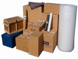 Stos kartonowych pudeł, skrzynia z drewna, rolka folii bąbelkowej oraz materiały do pakowania, przygotowane do transportu.