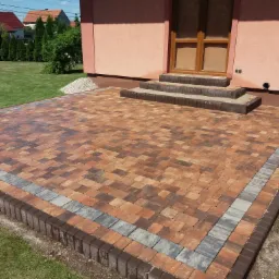 Wykończony taras z kostki brukowej w odcieniach brązu i szarości, z obrzeżem z ciemnobrązowych kostek, prowadzący do wejścia do domu z trzema schodkami, na tle zielonego trawnika i elewacji budynku.