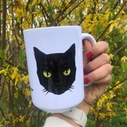 Studio Graficzne AGATA SCHLEISS - Biały kubek z grafiką czarnego kota w stylu low poly, trzymany w dłoni z czerwonym manicure na tle kwitnącej forsycji. Detal: napis w dolnej części kubka.