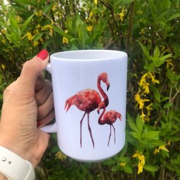 Studio Graficzne AGATA SCHLEISS - Biały kubek z poligonalnym wzorem flamingów trzymany w dłoni na tle kwitnącego krzewu. Widoczny fragment zegarka na pasku i czerwony lakier na paznokciach.