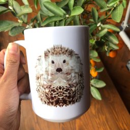 Studio Graficzne AGATA SCHLEISS - Biały kubek z geometrycznym wzorem jeża, trzymany w dłoni, na tle drewnianego stołu i rośliny cytrusowej z małymi pomarańczami.