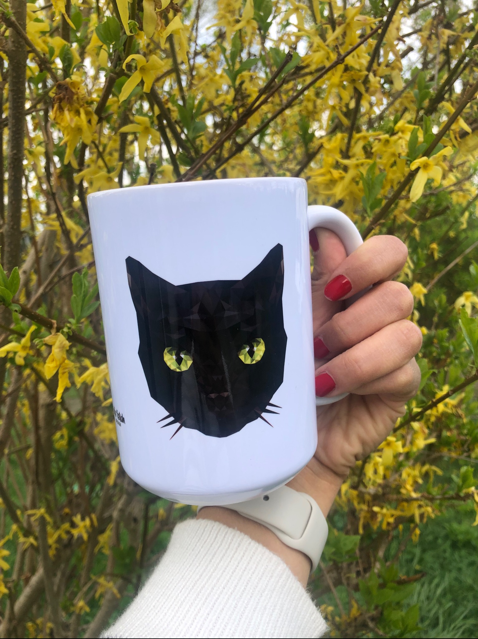 Biały kubek z grafiką czarnego kota w stylu low poly, trzymany w dłoni z czerwonym manicure na tle kwitnącej forsycji. Detal: napis w dolnej części kubka.