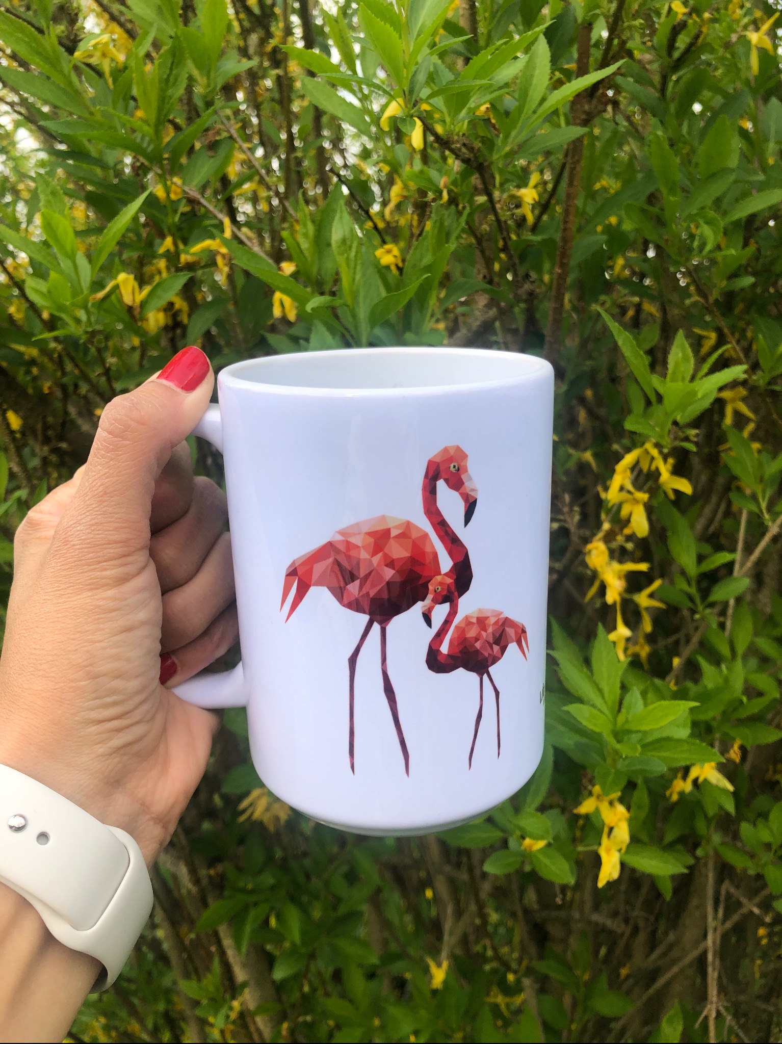 Biały kubek z poligonalnym wzorem flamingów trzymany w dłoni na tle kwitnącego krzewu. Widoczny fragment zegarka na pasku i czerwony lakier na paznokciach.