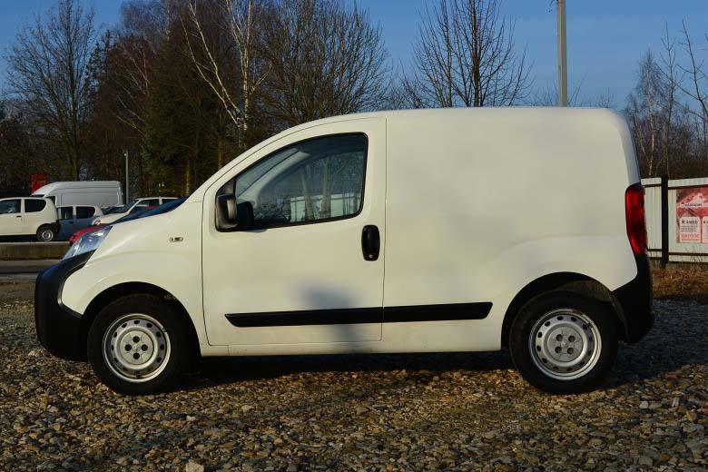 Biały samochód dostawczy z widocznymi kołpakami na kołach, zaparkowany na żwirowym parkingu w słoneczny dzień.