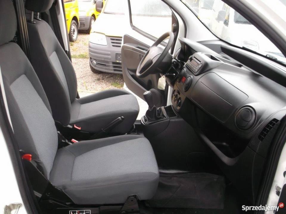 Wnętrze białego samochodu dostawczego z szarymi fotelami, widok z otwartych drzwi na deskę rozdzielczą i dźwignię zmiany biegów.