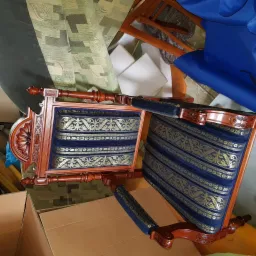 Dwa odrestaurowane krzesła z drewnianą ramą i niebieską tapicerką w złote wzory, widoczne w otoczeniu materiałów tapicerskich i kartonu.