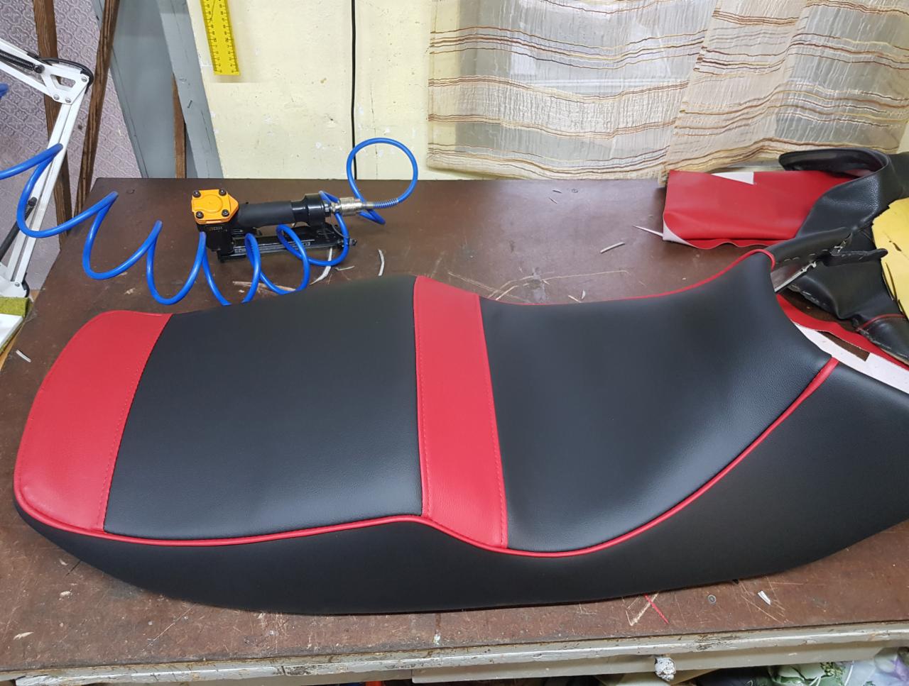 Nowe, czarno-czerwone obicie siedziska motocyklowego na stole warsztatowym, widoczny zszywacz pneumatyczny i resztki materiałów.