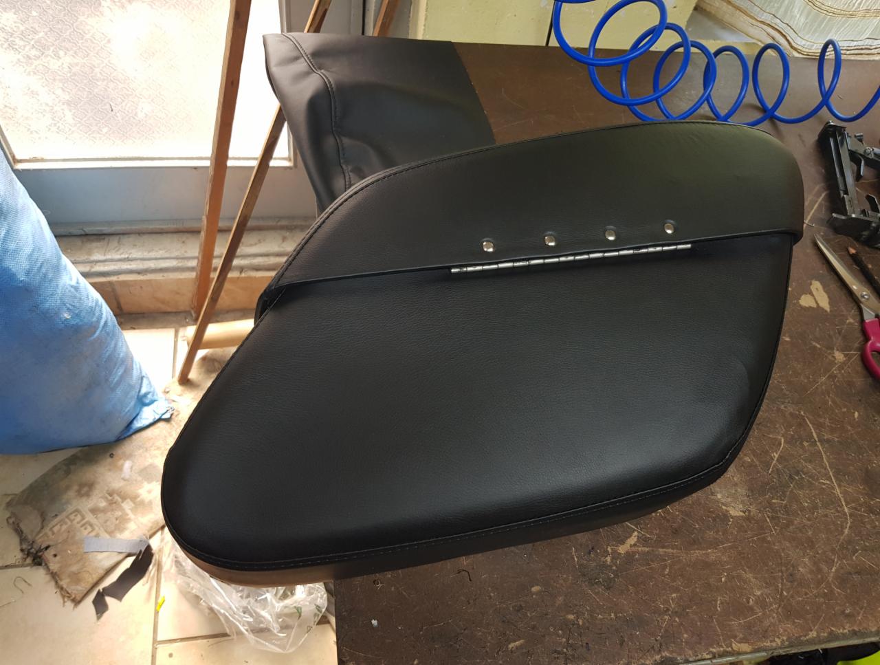 Czarna sakwa motocyklowa z metalowymi zawiasami i nitami, leżąca na stole warsztatowym obok narzędzi.