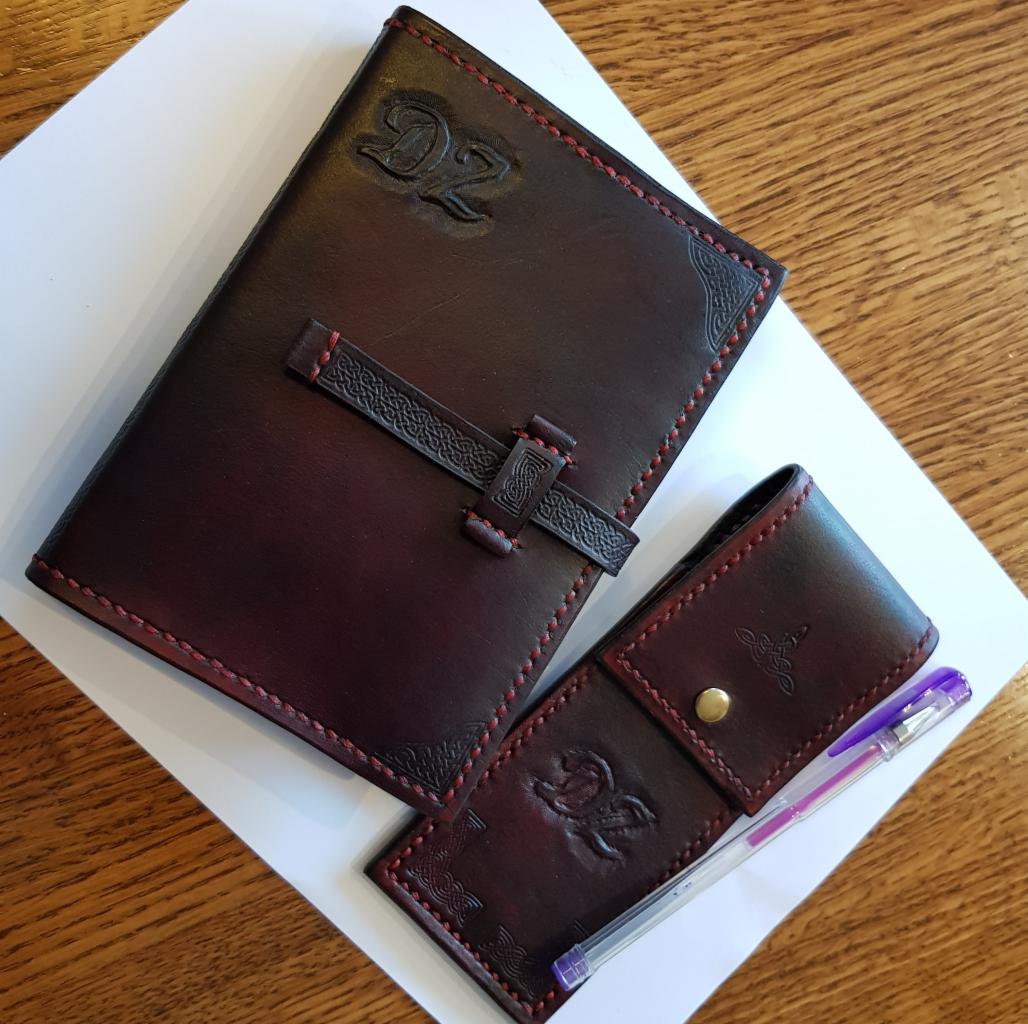 Ręcznie wykonany, skórzany notatnik w kolorze burgundowym z ozdobnym zapięciem i inicjałami 'DZ' oraz dopasowane etui na długopis, oba wykończone czerwoną nicią, leżące na białej kartce...