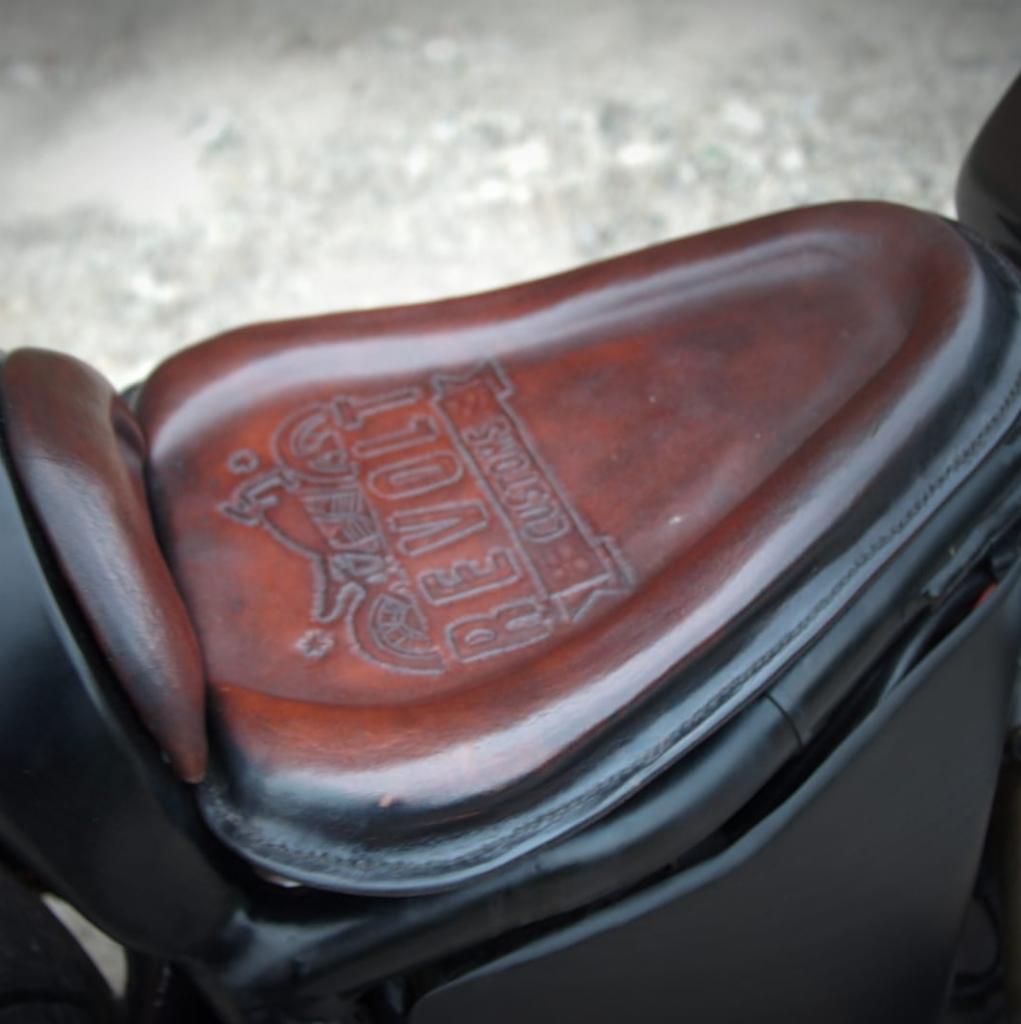 Brązowe, skórzane siedzenie motocyklowe z wytłoczonym logo 'REVOLT CUSTOMS', zamontowane na czarnym motocyklu.