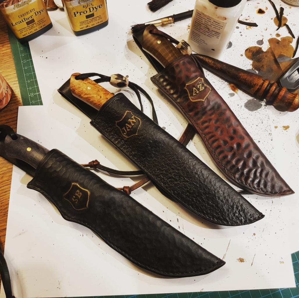 Trzy ręcznie wykonane skórzane pochwy na noże z wytłoczonymi inicjałami (SB, JAN, AZ) leżą na stole warsztatowym obok barwników do skóry i narzędzi.
