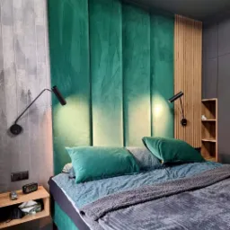 Sypialnia z panelem tapicerowanym w odcieniach zieleni za łóżkiem, uzupełniona o drewniane lamele i minimalistyczne lampki nocne.