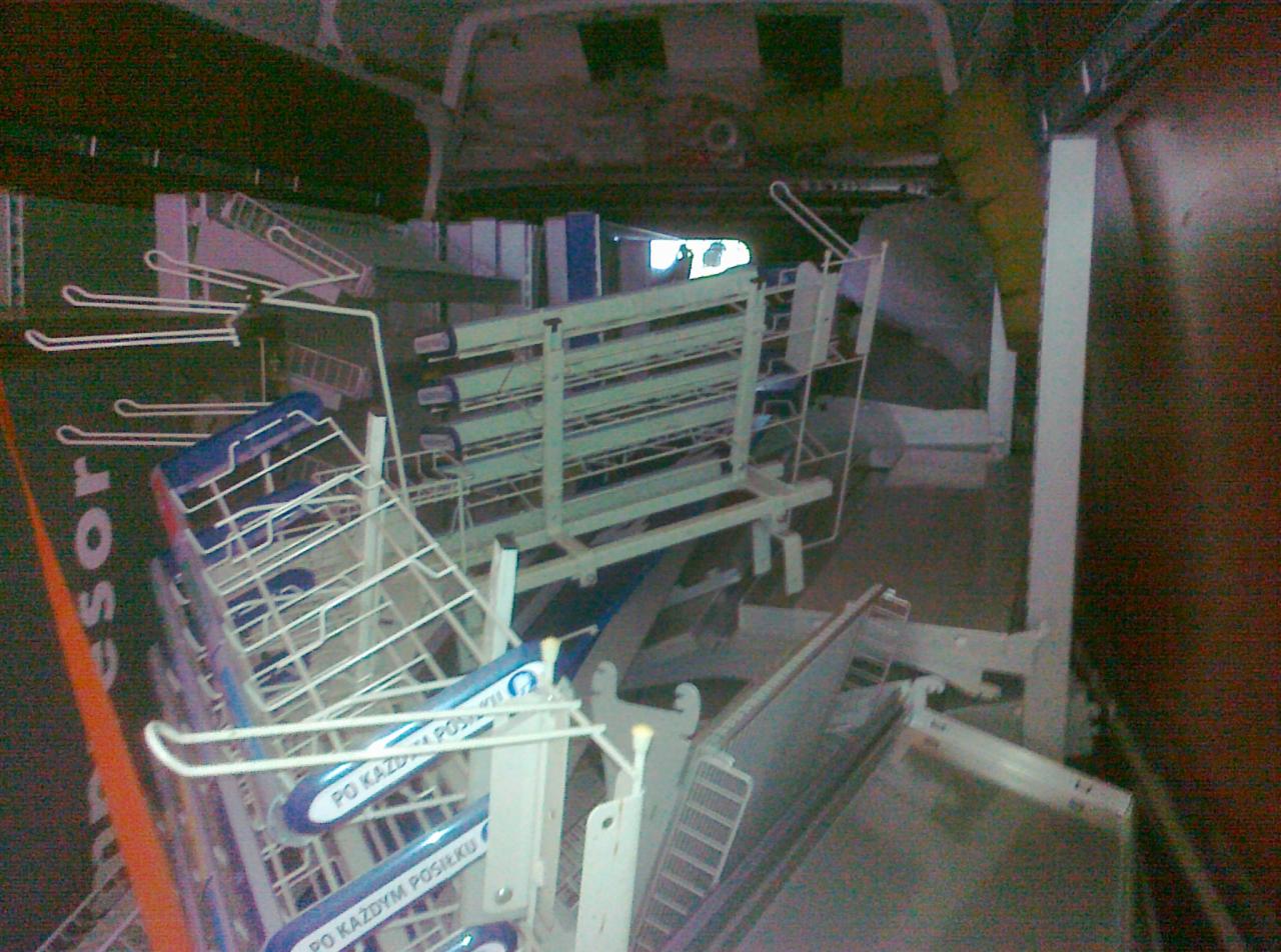 Wnętrze pojazdu transportowego załadowane metalowymi regałami i wózkami z niebieskimi uchwytami, widoczne napisy 'PO KAŻDYM POSIŁKU' na uchwytach.