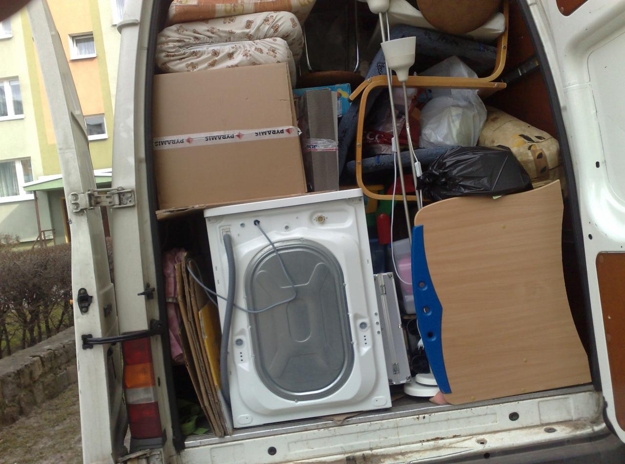 Wnętrze furgonetki załadowane rzeczami do transportu, widoczna pralka, karton z taśmą 'PYRAMIS', lampa stojąca i inne przedmioty.