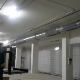 Długi, segmentowy kanał wentylacyjny z perforowanymi elementami, zamontowany pod sufitem w pomieszczeniu wyłożonym białymi płytkami, widoczny podnośnik nożycowy.